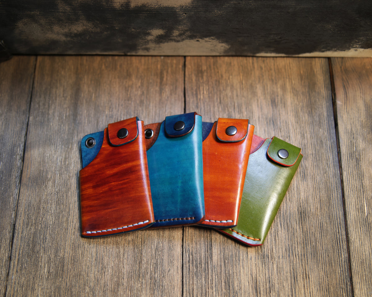 Four hand-dyed Scout mini-trucker wallets on a rustic wood surface, including two Bourbon & Blues dye variations, Deep Blue & Brown, and Army Green & Orange.