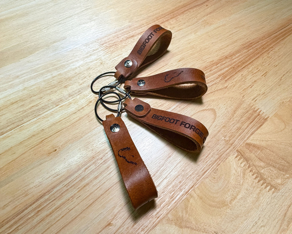 Leather EDC keychains in bourbon with engraved Bigfoot Forge branding and steel cable rings laying flat