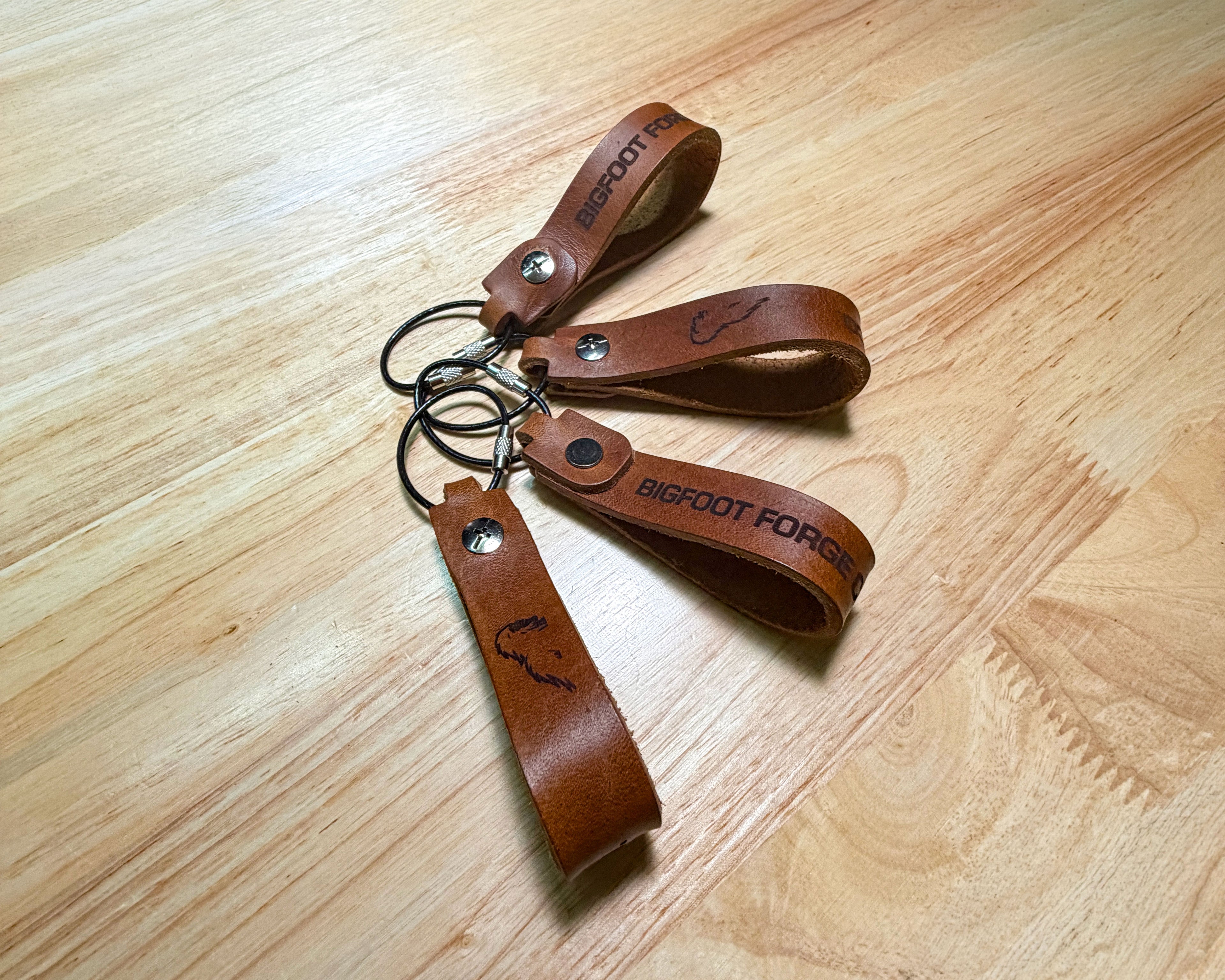 Leather EDC keychains in bourbon with engraved Bigfoot Forge branding and steel cable rings laying flat