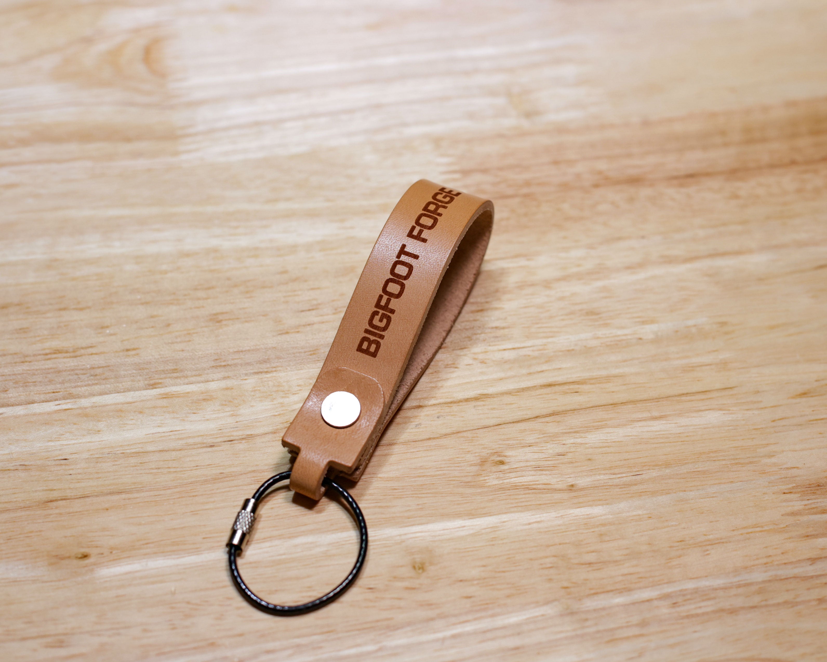 Veg tan leather keychain with engraved Bigfoot Forge text and coated stainless steel cable key ring on wood surface