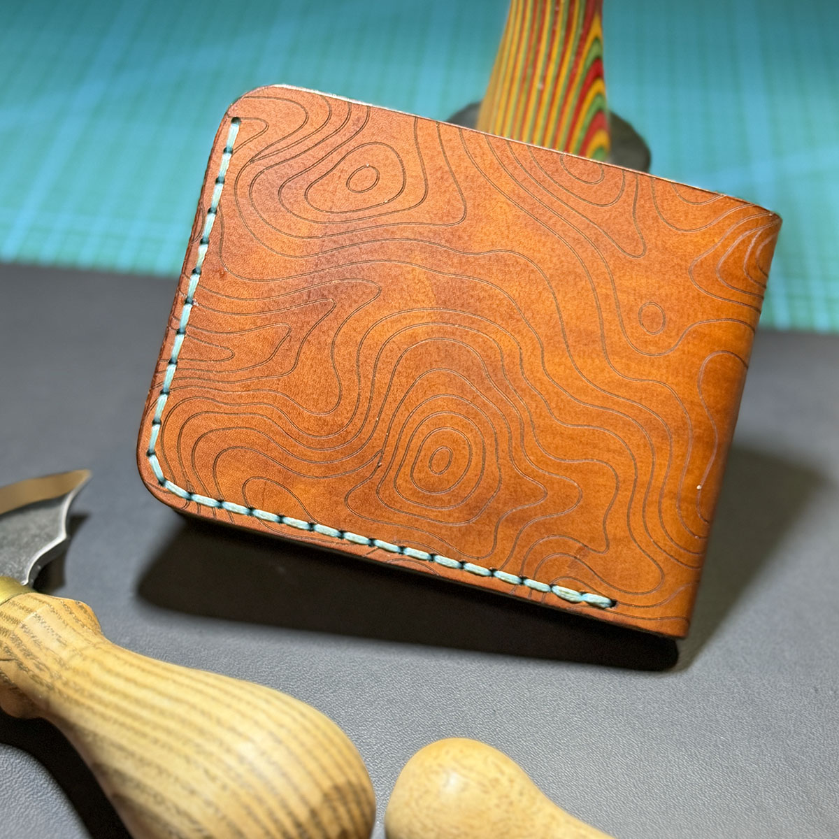Bourbon topo leather wallet with blue hand stitching, photographed beside leatherworking tools.