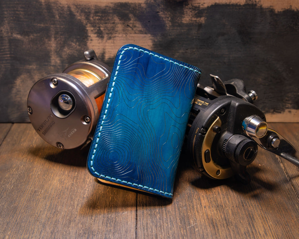 Angled exterior of the Forgehand Field Deep Blue Water wallet showing the hand-dyed blue gradient and topographic engraving.
