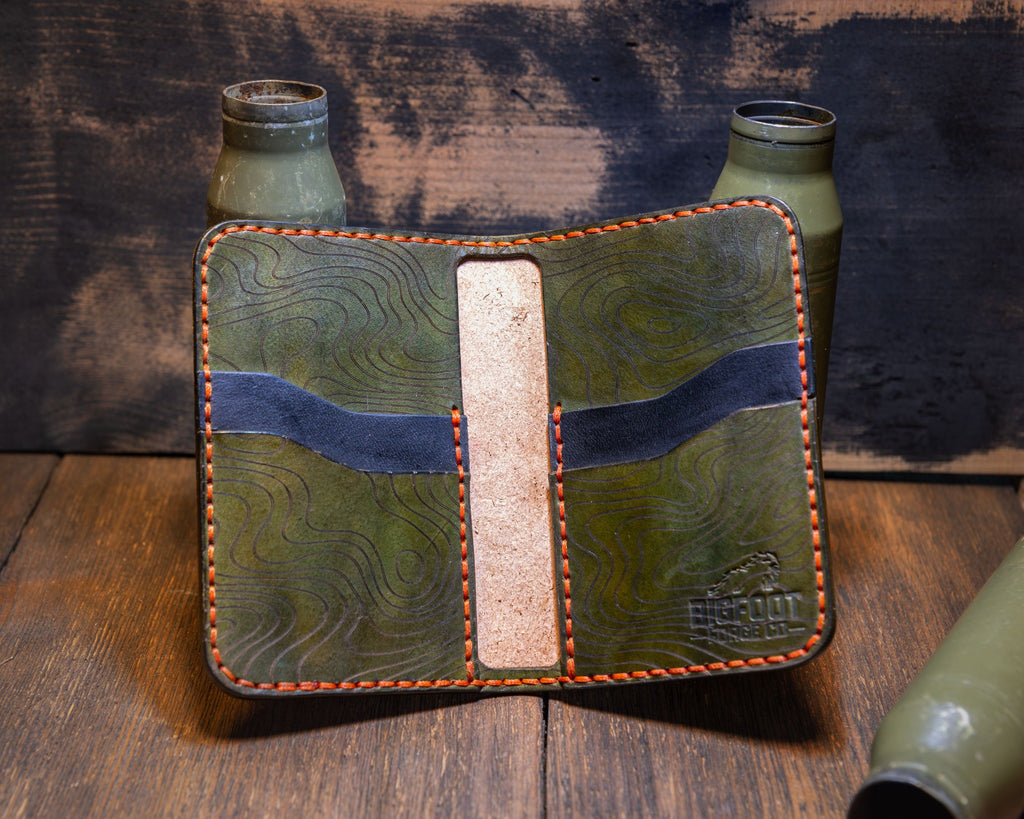 Open Woodland Camo vertical bifold with green topographic card pockets, blue accents and a natural leather center strip.